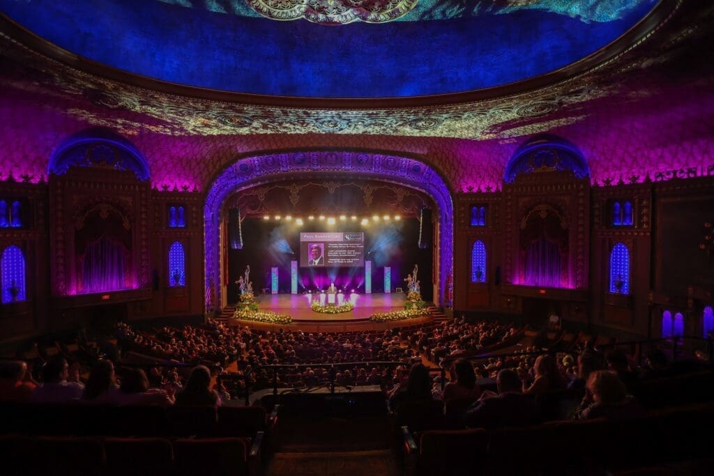 A vibrant theater stage lit in purple and blue with an audience watching.
