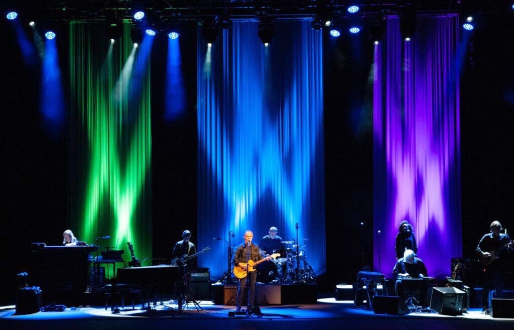 A band performs on stage with colorful blue, green, and purple lighting.