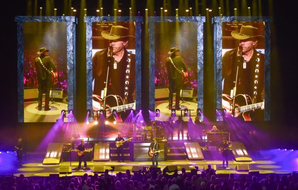 A musician performs on stage with multiple large screens behind him.