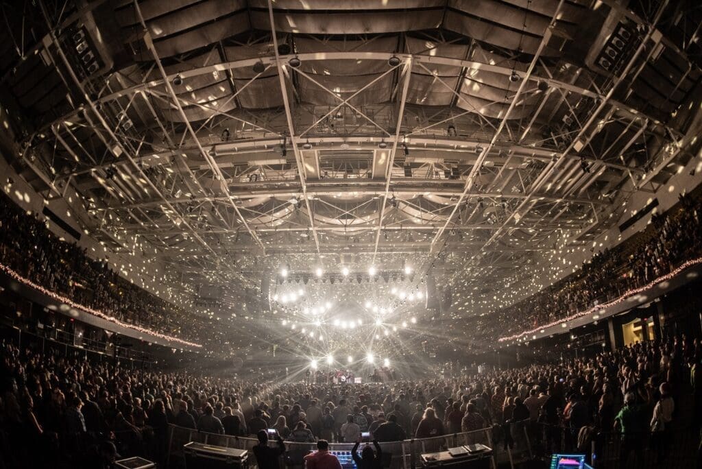 A packed arena shines under bright stage lights during a live concert.