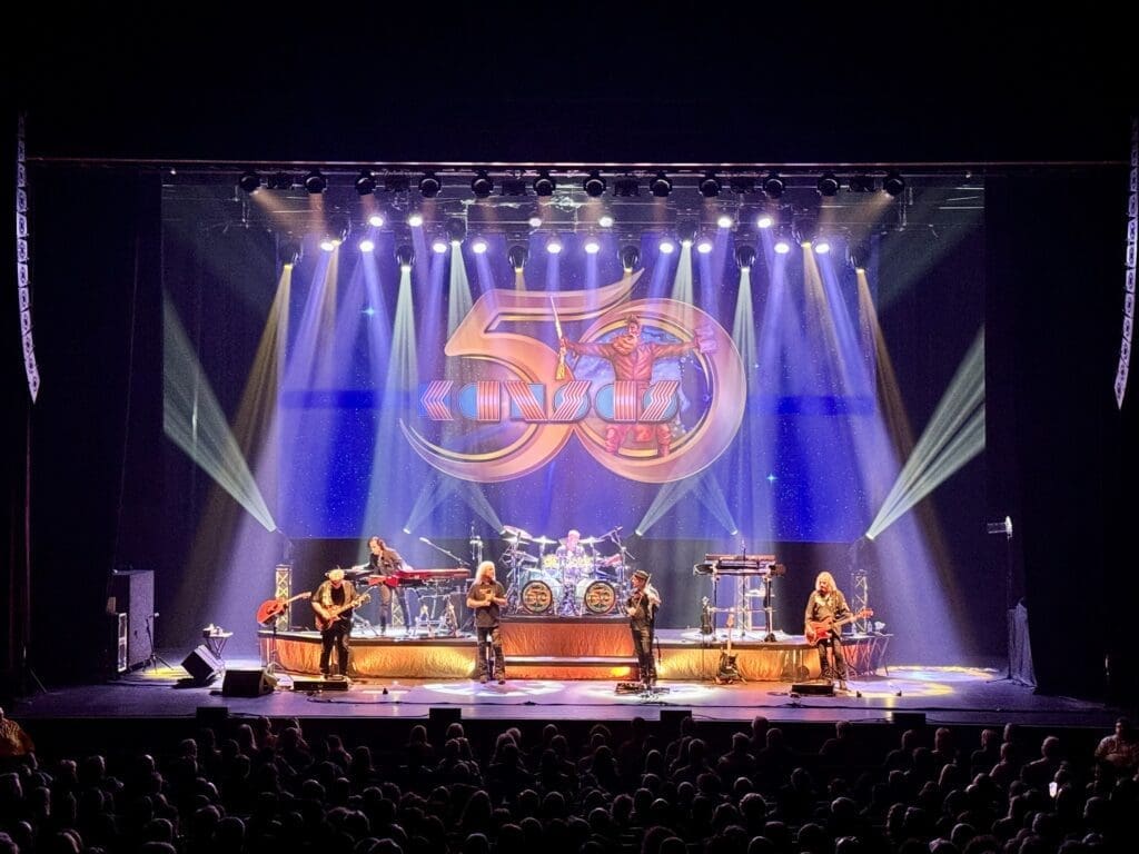 A live band performs on stage with vibrant lighting and a large logo backdrop.