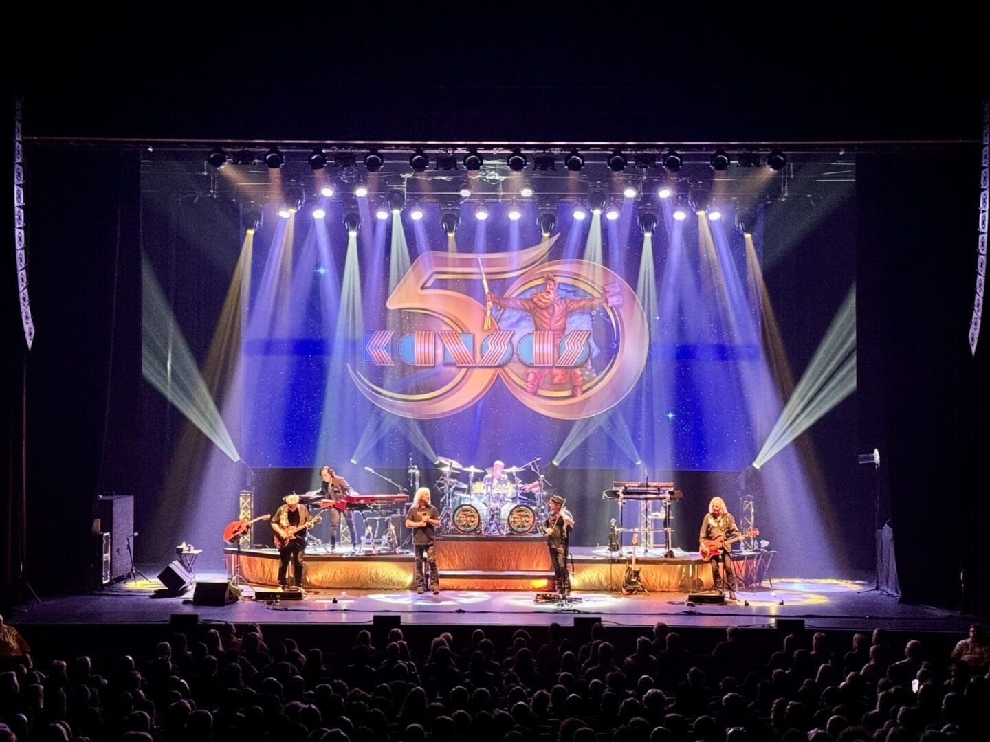 A live band performs on stage with vibrant lighting and a large logo backdrop.