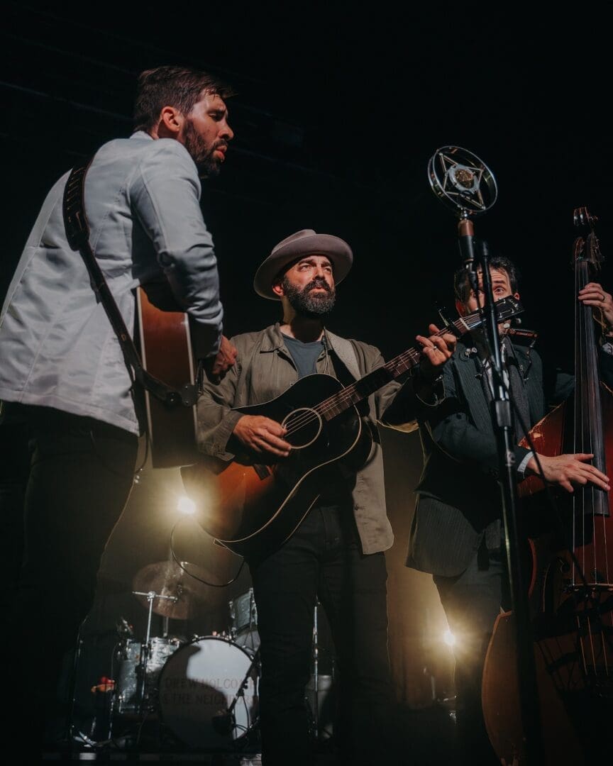A trio of musicians performing under stage lights with guitars and a microphone.