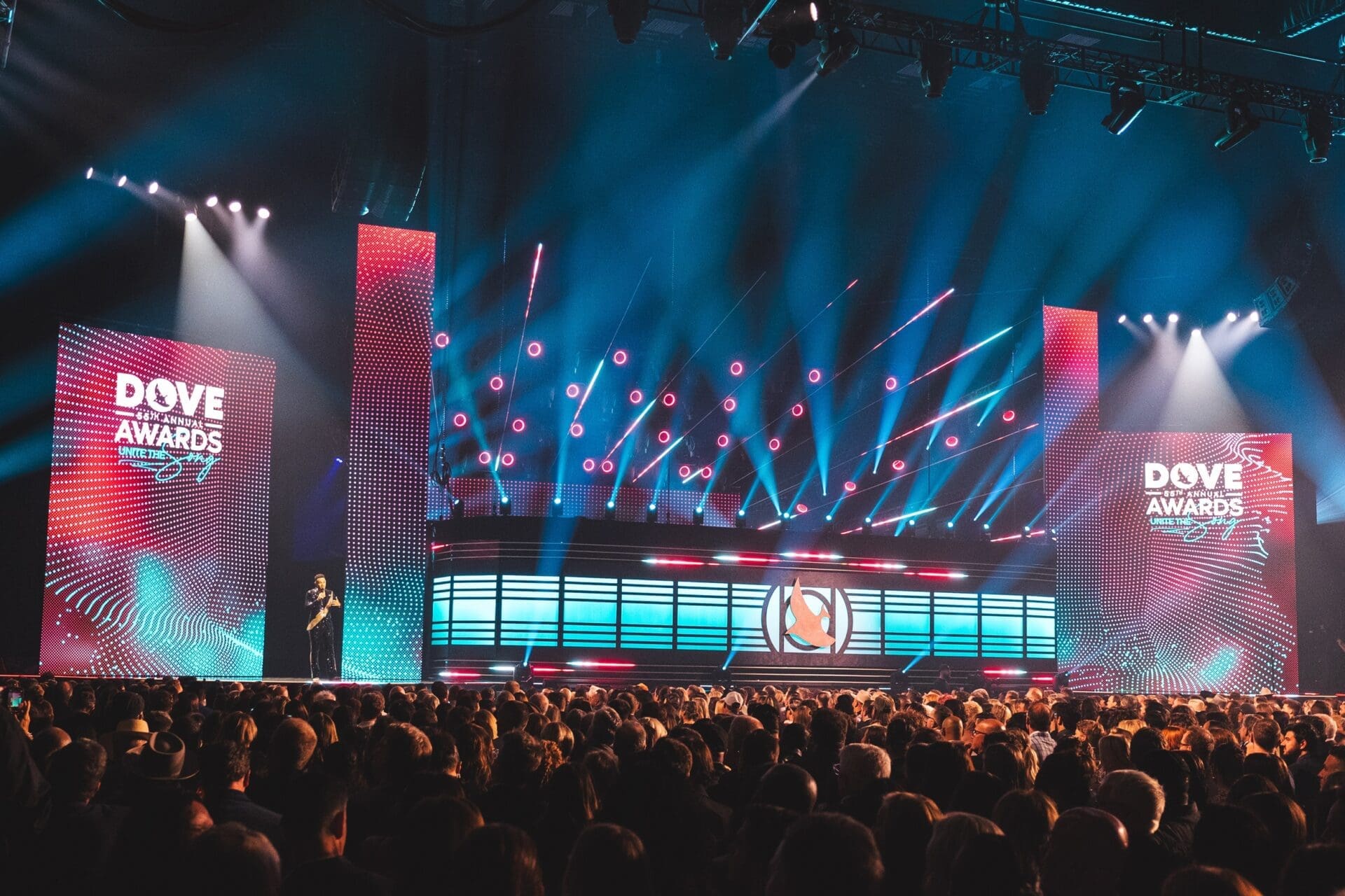 A large crowd watches a vibrant light show on a stage with illuminated screens.