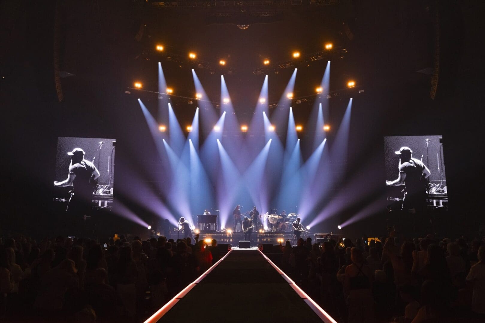 A band performs on a brightly lit stage with large screens and an enthusiastic audience.