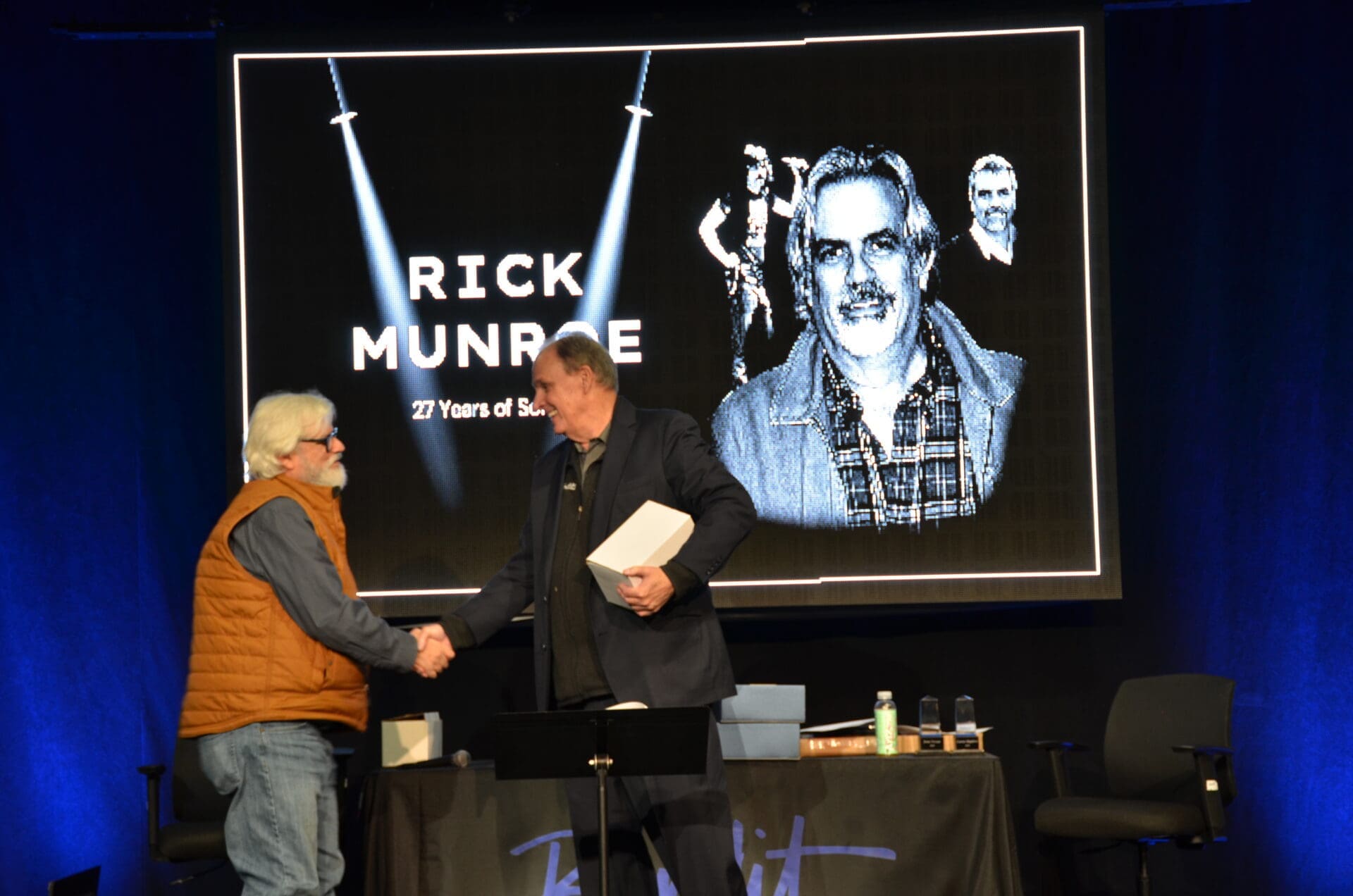 Two men shaking hands in front of a Rick Munroe tribute screen.