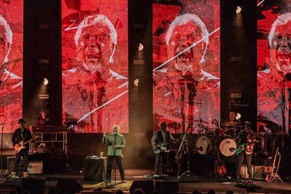 Band performs live on stage with large red visual screens behind.