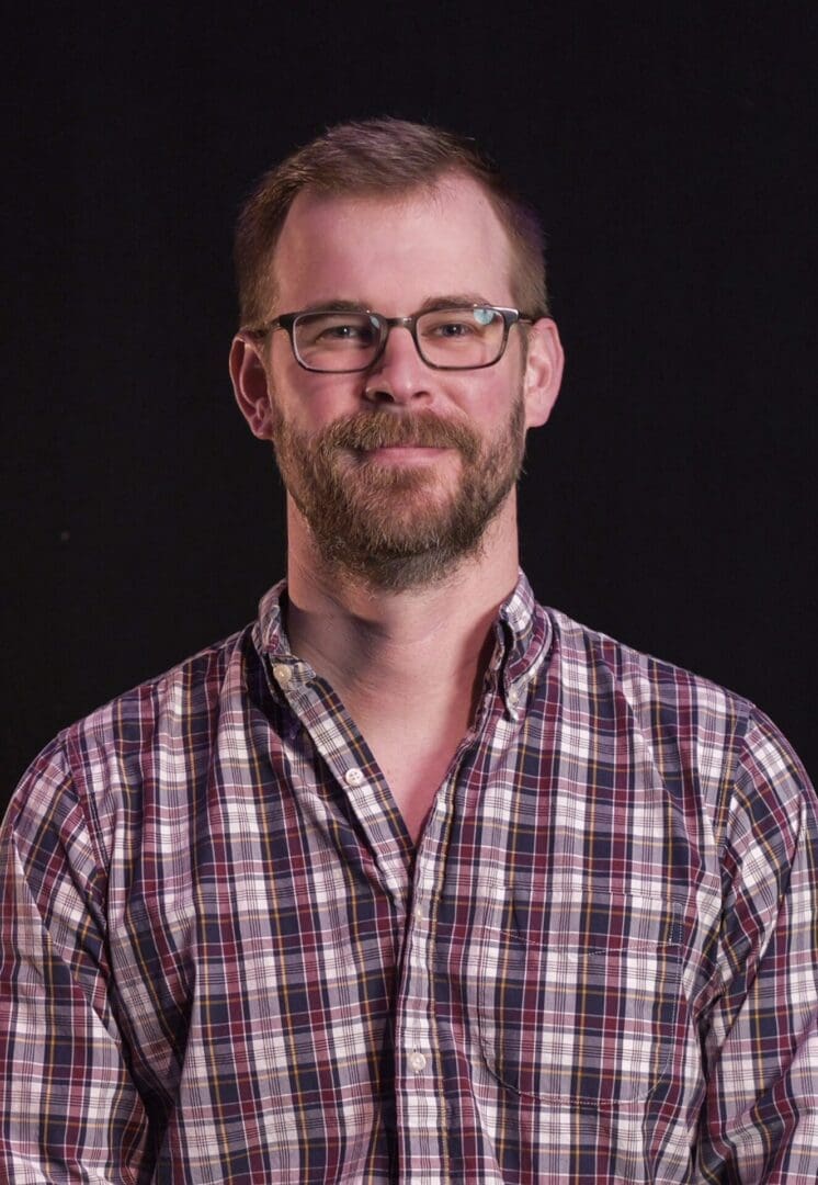 Smiling man wearing glasses and a plaid shirt against a dark background.