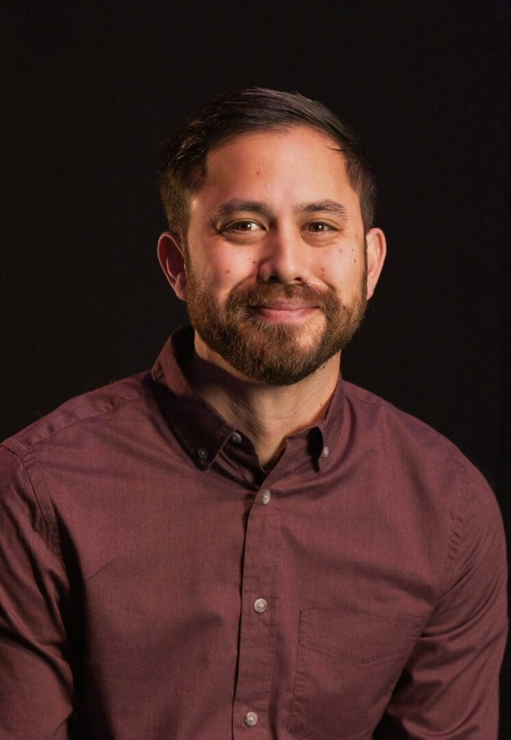 Portrait of a smiling man in a maroon shirt against a dark background.
