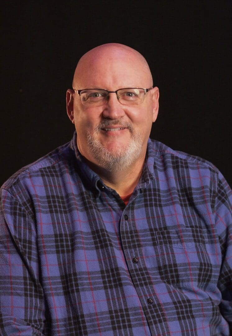 Smiling man with glasses and a plaid shirt against a dark background.