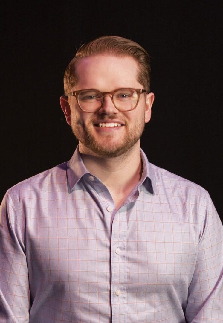 Man with glasses and beard smiling in a light purple shirt.