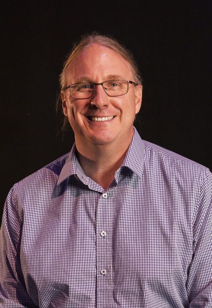 Smiling man wearing glasses and a checkered shirt against a black background.