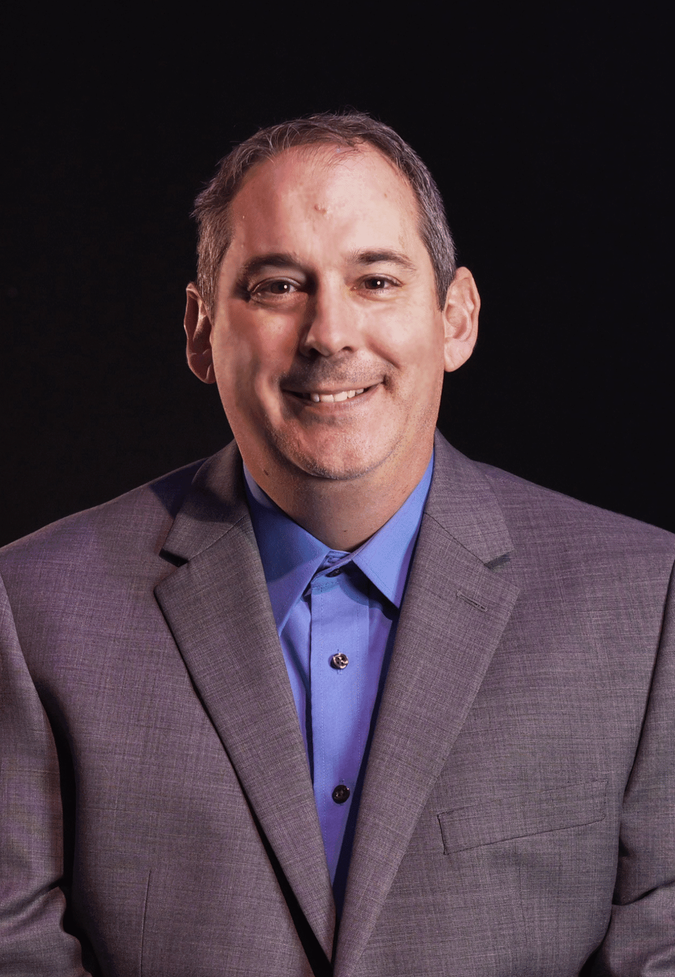 Smiling man in a grey suit and blue shirt against a black background.
