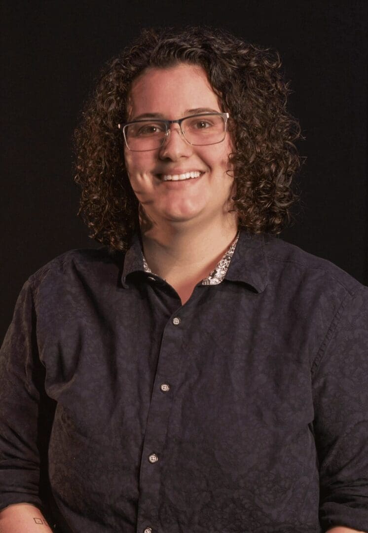 Smiling person with curly hair and glasses in a dark shirt.