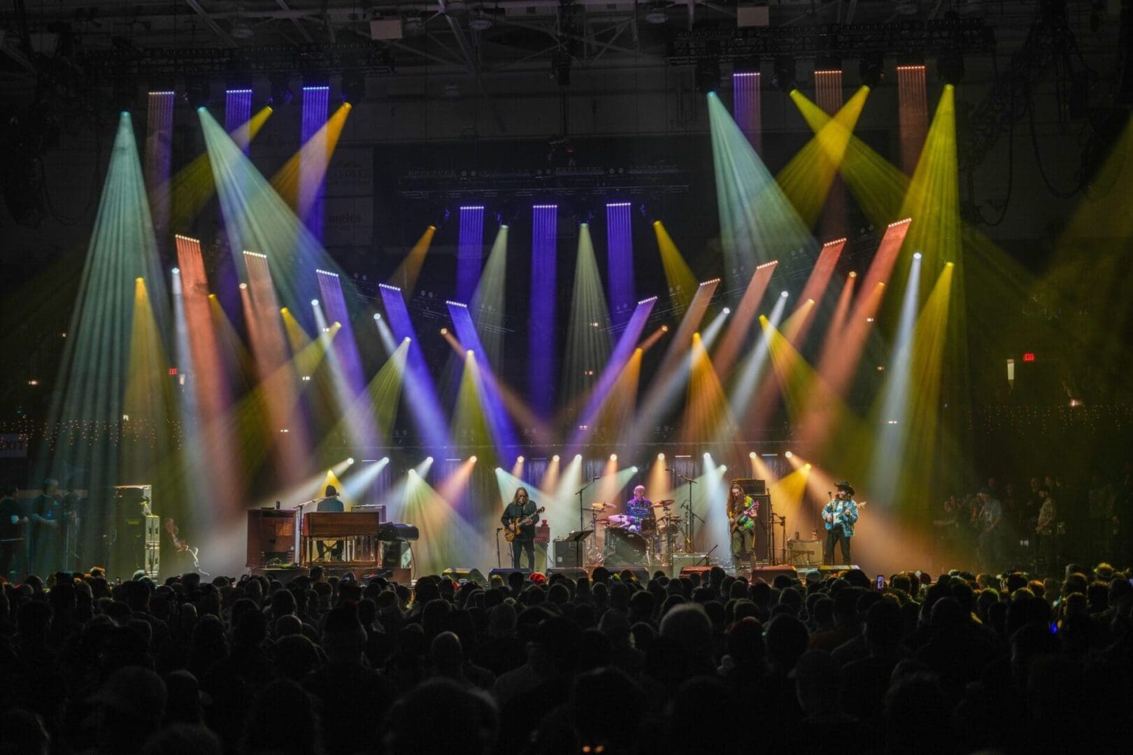 Colorful stage lights illuminate a live concert with performers and an enthusiastic crowd.