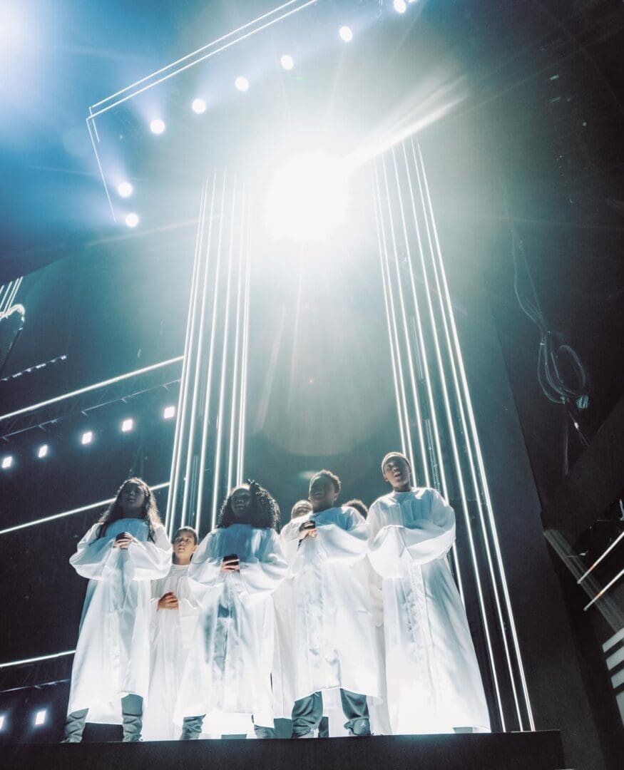 Performers in white costumes illuminated by bright stage lights.