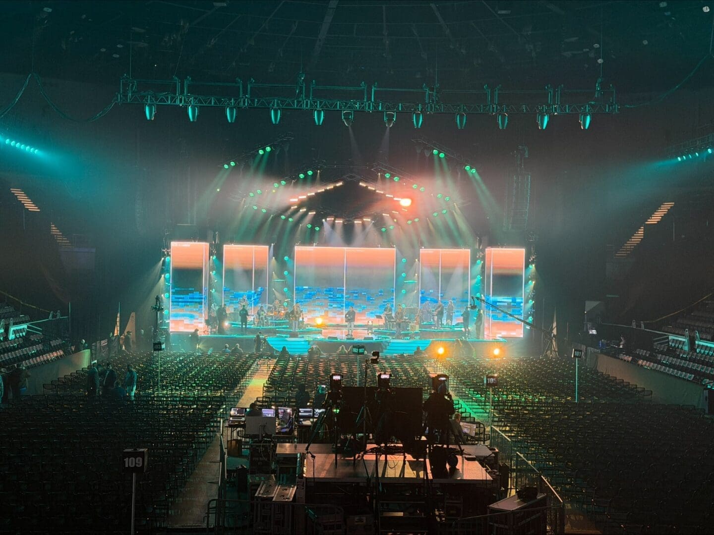 Concert stage illuminated with vibrant lights and large screens in a packed arena.