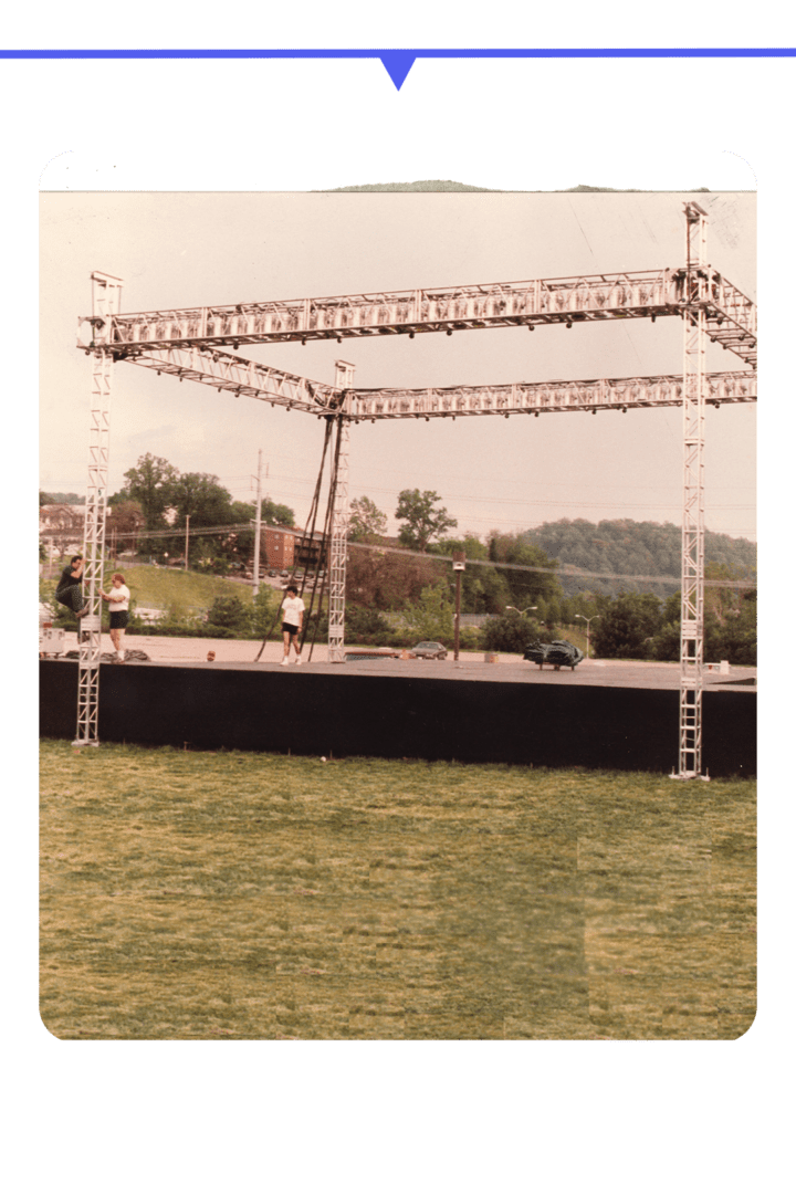 An outdoor stage setup with metal scaffolding and speakers on a grassy field.