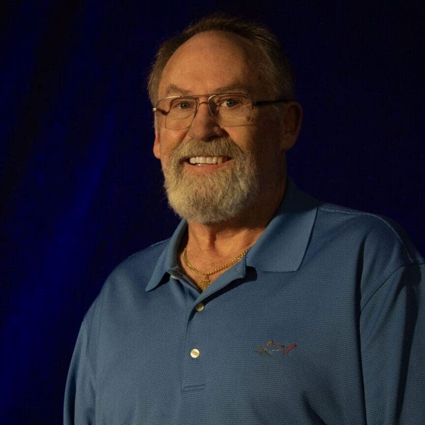 Smiling elderly man with glasses and beard in a blue polo shirt.