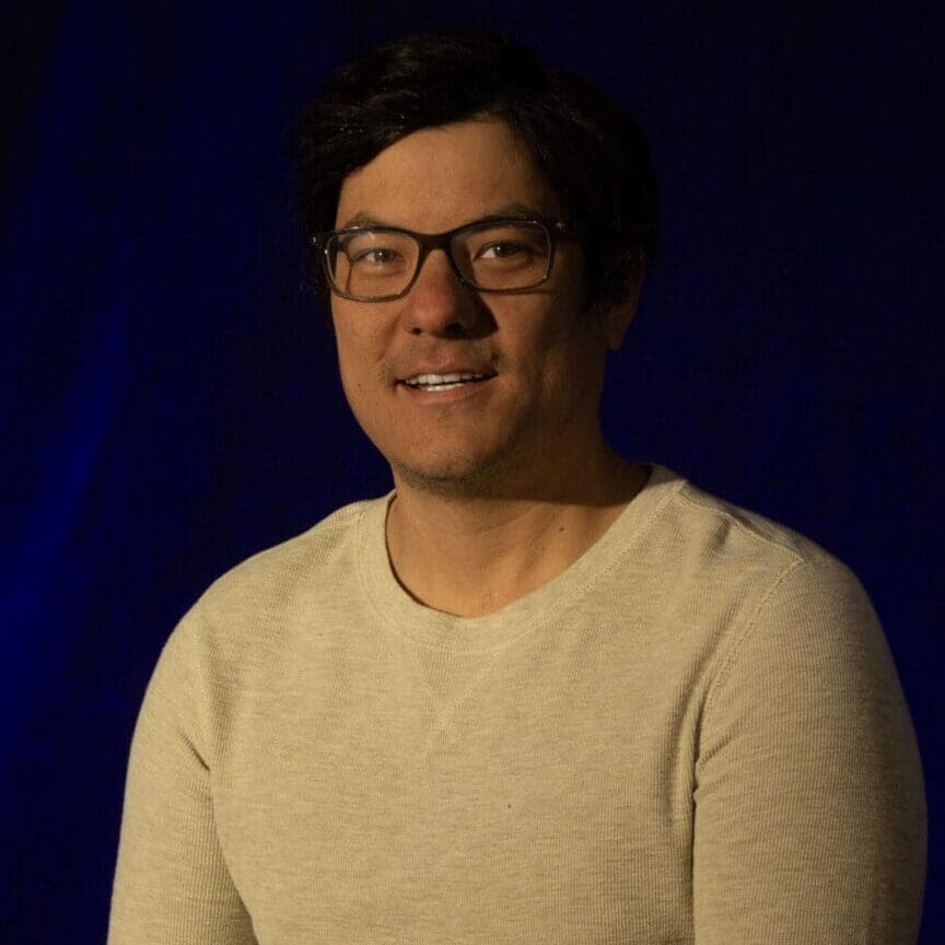 Man with glasses smiling against a dark background.
