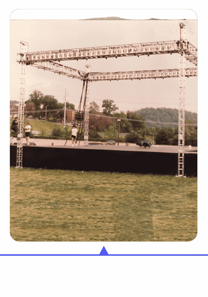 Outdoor stage setup with people preparing under a cloudy sky.